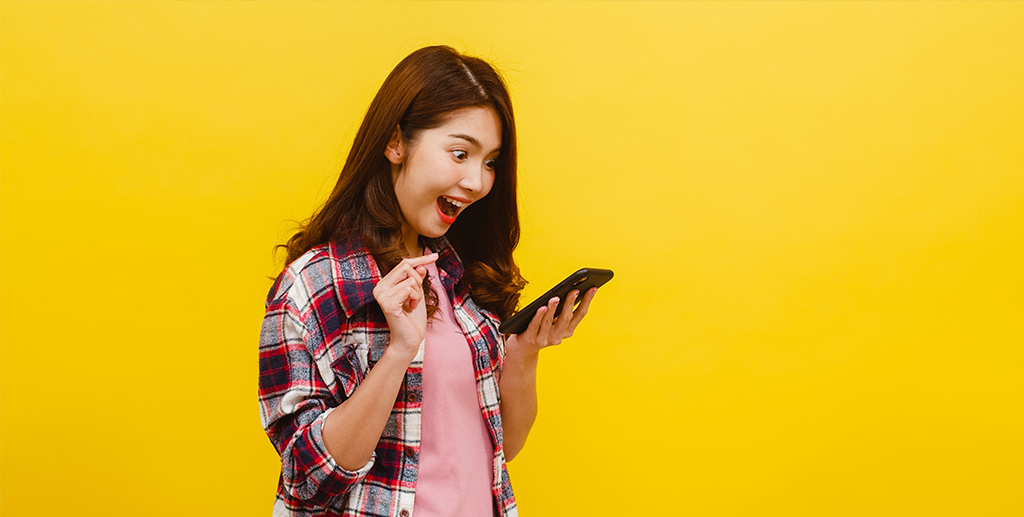 portrait-surprised-asian-female-using-mobile-phone-with-positive-expression-dressed-casual-clothing-looking-camera-yellow-wall-happy-adorable-glad-woman-rejoices-success
